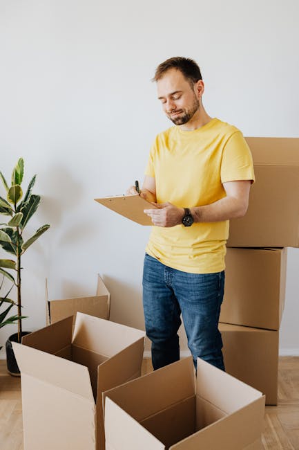 A man wearing a yellow t-shirt and blue jeans stands indoors, holding a clipboard and pen, seemingly preparing for a home relocation or moving process. Around him are several open cardboard boxes, som