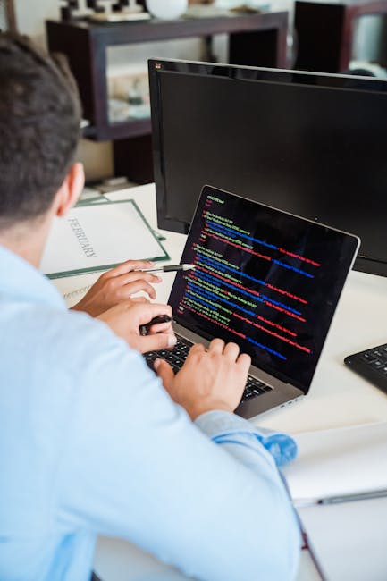 A person seated at a desk in an indoor office environment, viewed from behind, working on a laptop with a screen displaying lines of code in various colors. The desk includes a black keyboard, a black